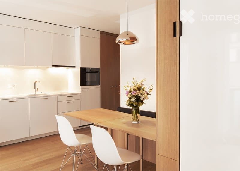 Modern kitchen with white cabinets, a wooden dining table, white chairs, a vase with flowers, and a hanging pendant light.