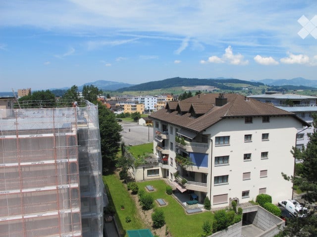 Multi-story residential building with balconies, surrounded by greenery and a view of distant mountains.