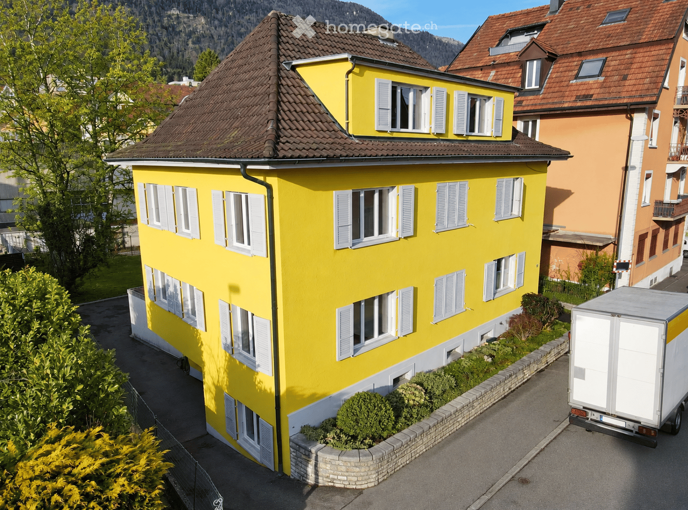 Bright yellow two-story house with multiple windows and white shutters, surrounded by greenery and a neighboring building.