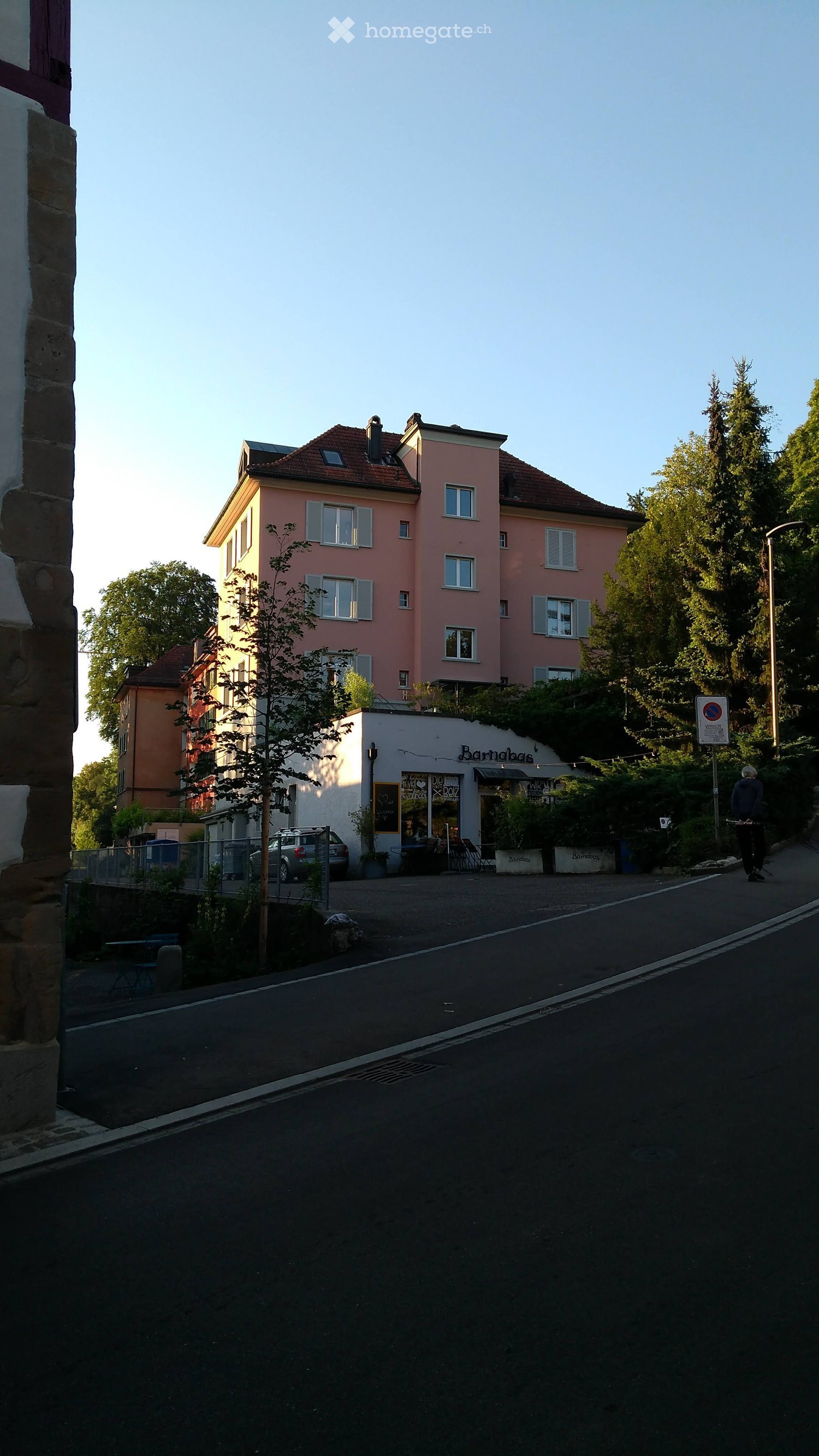 A multi-story residential building with pink facade and multiple windows, adjacent to a small shop with outdoor seating and a car parked nearby.