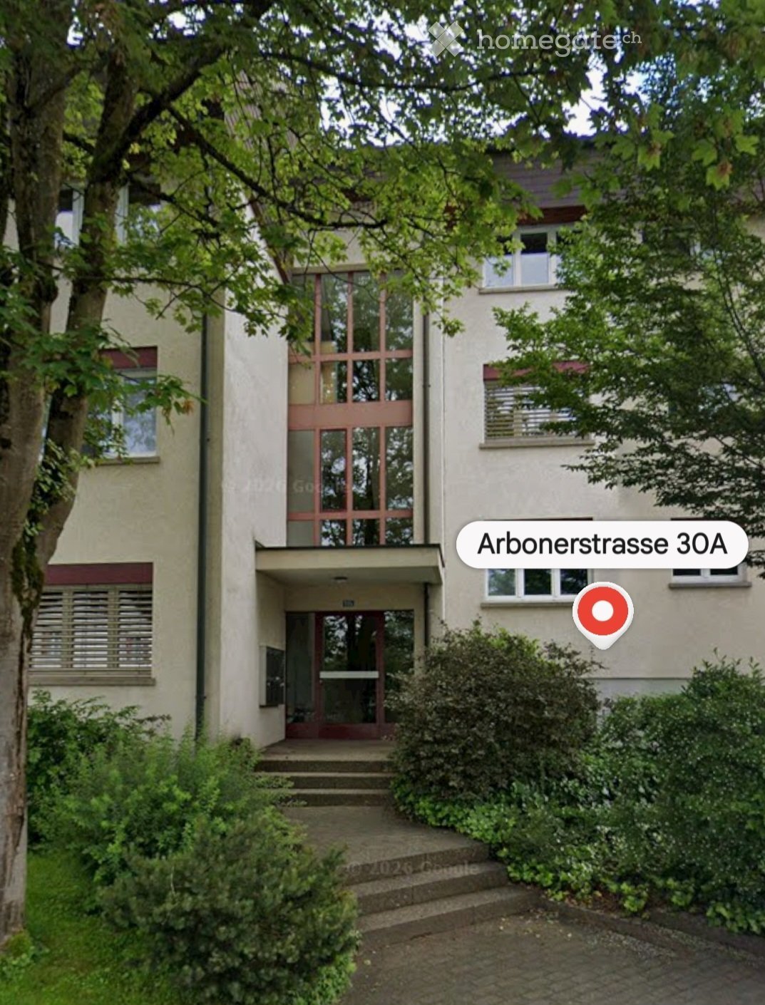 Two-story residential building with a modern facade, large windows, and a small entrance area with steps leading up to a glass door.