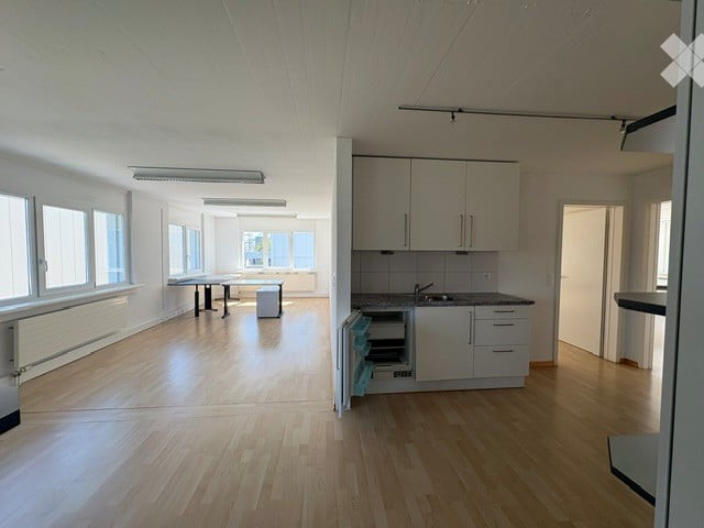 Bright, open-plan space with light wooden flooring, white walls, and large windows. Includes a small table with a stool, a fitted kitchen area with cabinets, a sink, and a faucet.