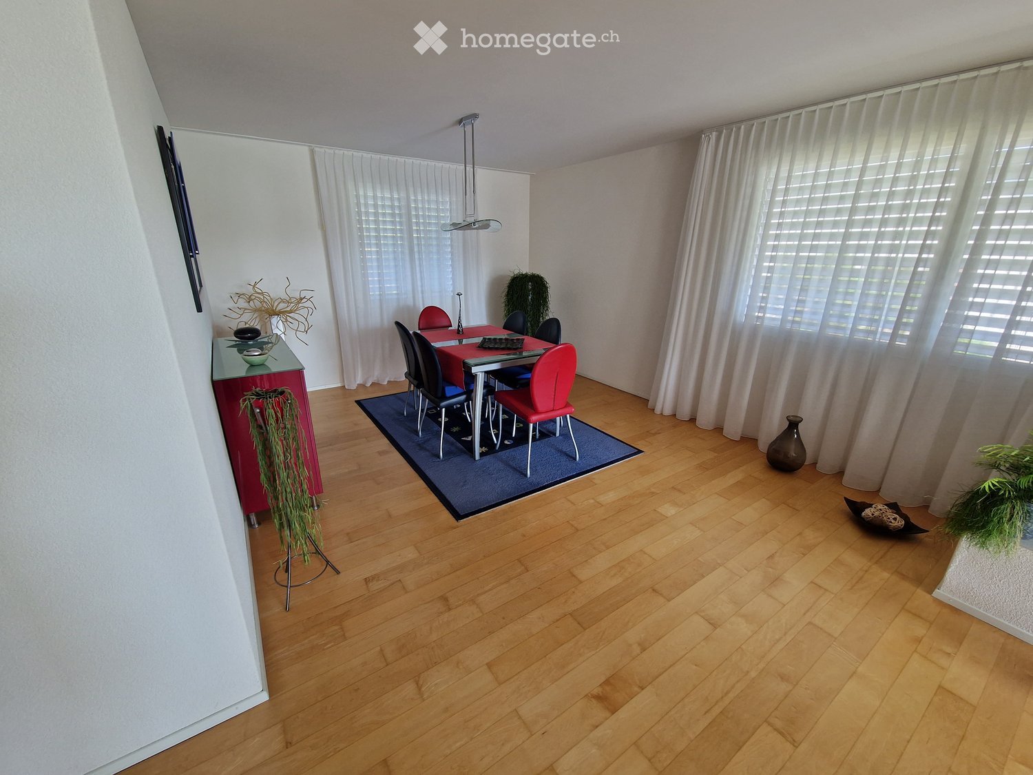 Dining room with wooden flooring, a red dining table surrounded by chairs, a sideboard, and decorative plants.