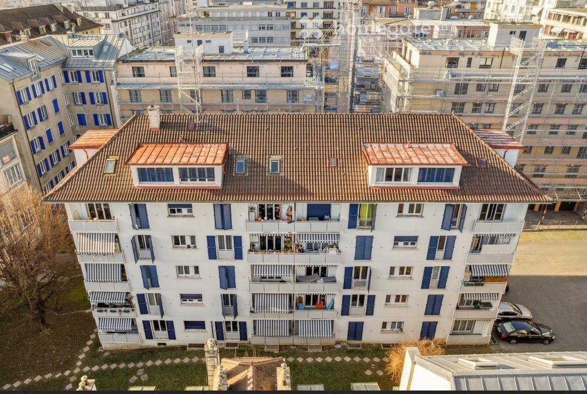 A multi-story residential building with white walls, blue shutters, and multiple balconies. The roof is tiled, and the surrounding area includes other buildings and parked cars.