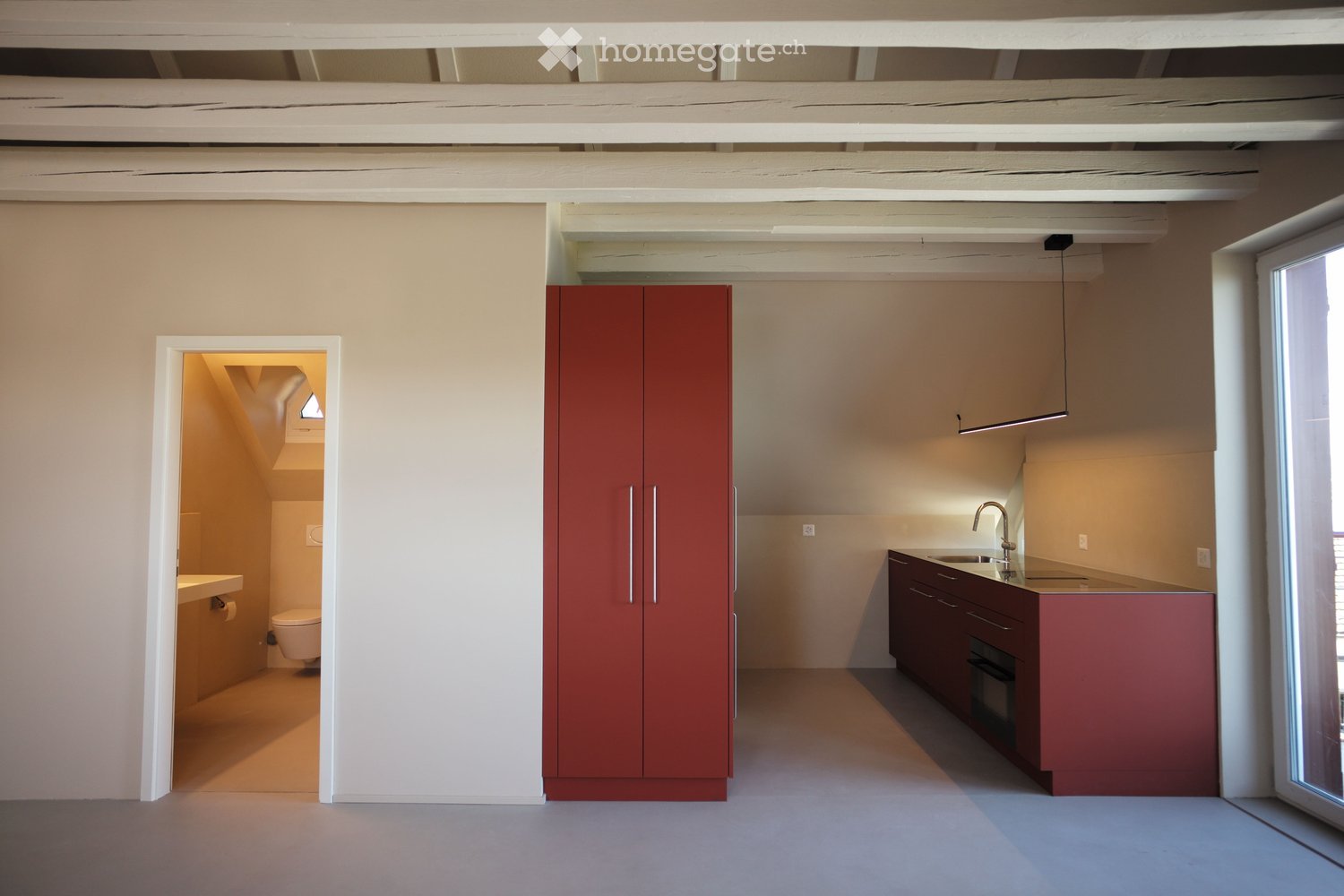 Modern kitchen with red cabinets, a sink, and an oven. An open doorway leads to a small bathroom with a toilet and sink.