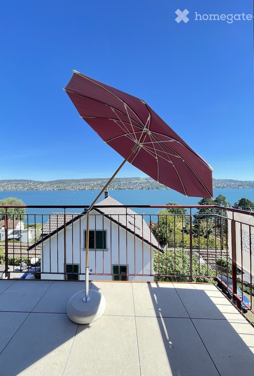 Balcony with a large umbrella, overlooking a lake and distant hills under a clear blue sky.