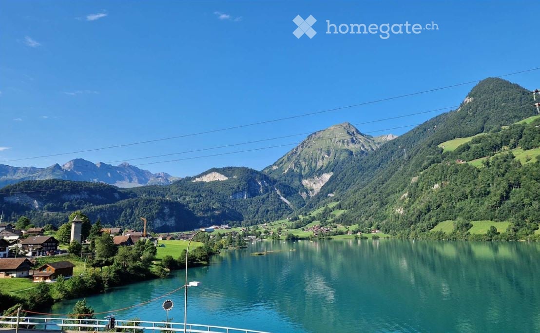 Scenic view of a lake surrounded by lush green hills and mountains, with small houses and a bridge visible in the foreground.