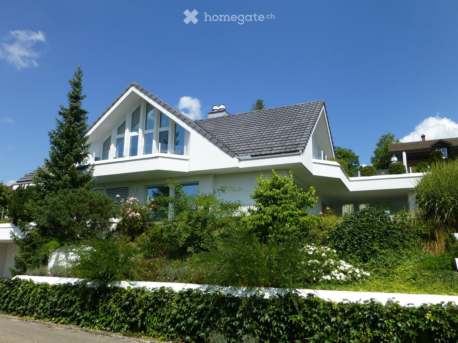 Modern two-story house with white exterior, large windows, and a sloped roof, surrounded by greenery and a well-maintained hedge.