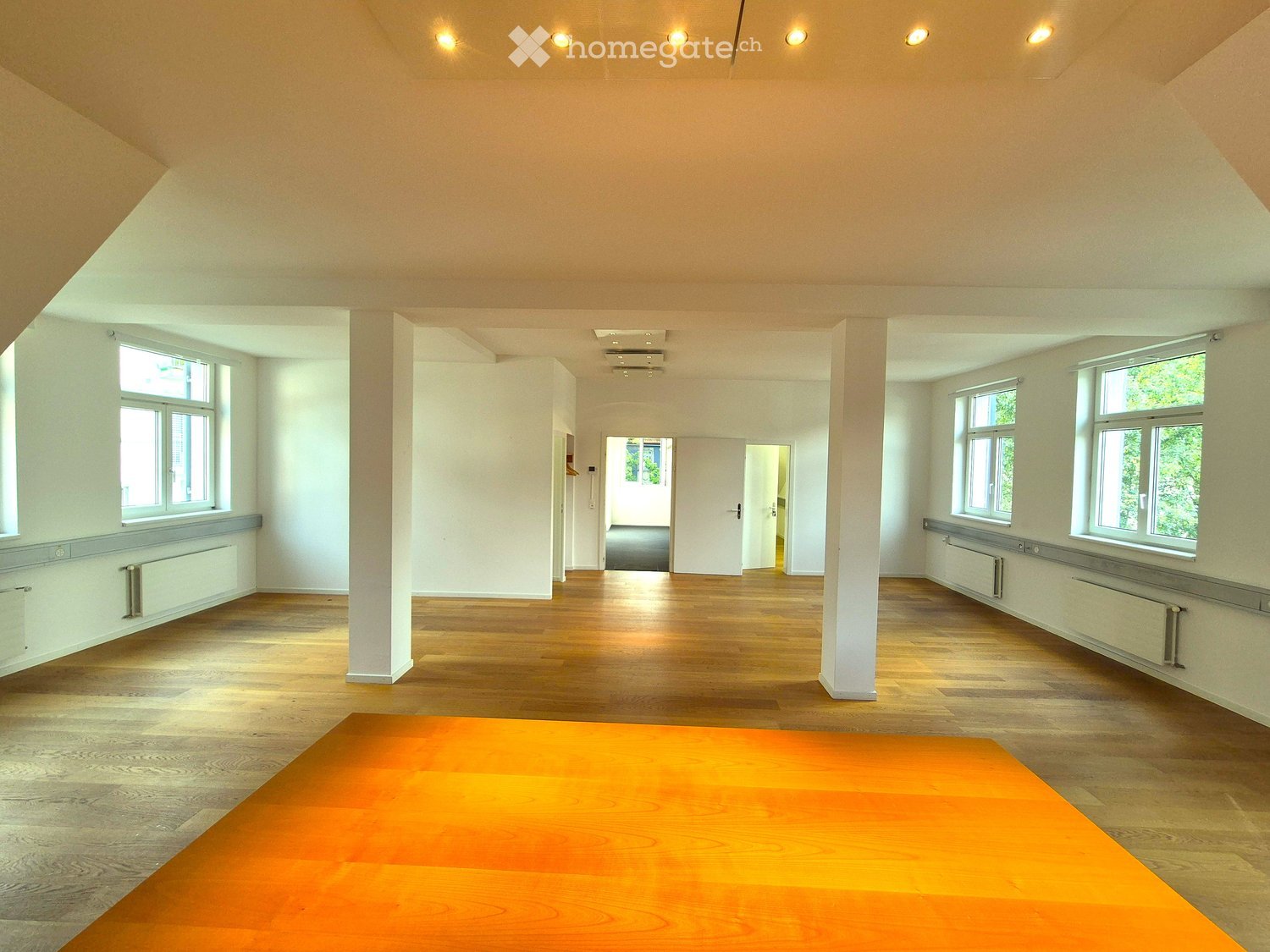 Empty room with wooden flooring, white walls, large windows, and a central column. No furniture or equipment present.
