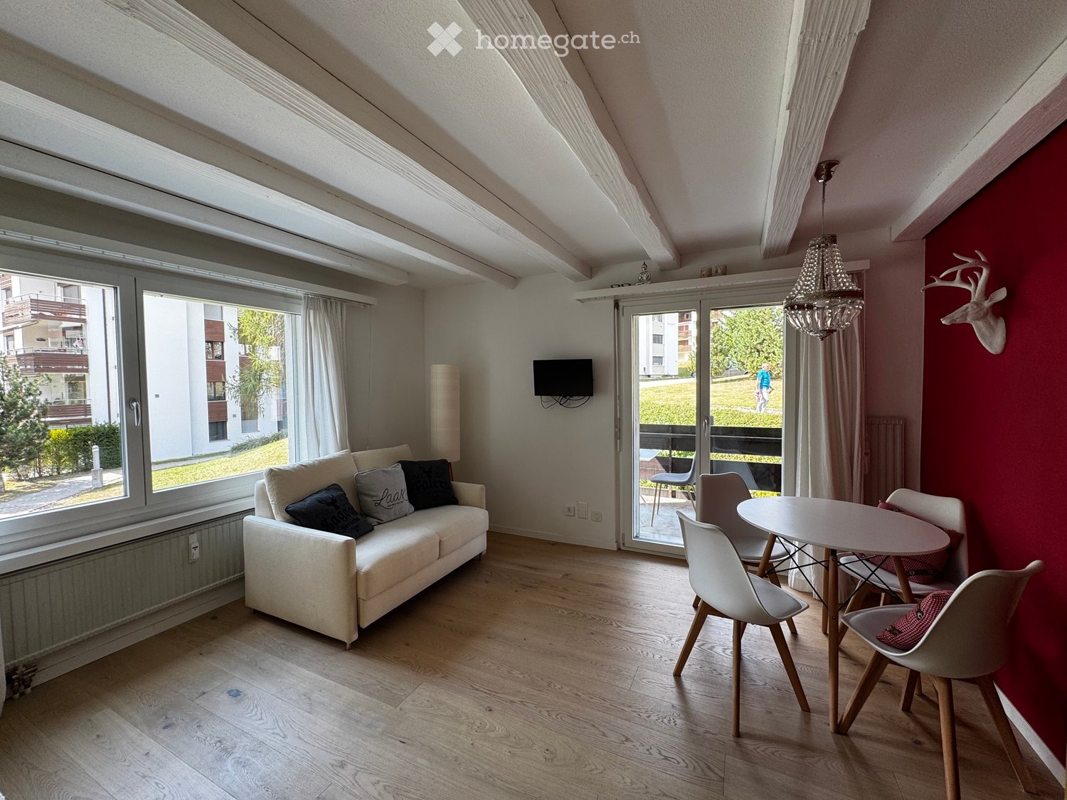 Bright living room with wooden flooring, large windows, a white sofa with decorative pillows, a dining table with chairs, and a wall-mounted TV. A chandelier hangs from the sloped ceiling.