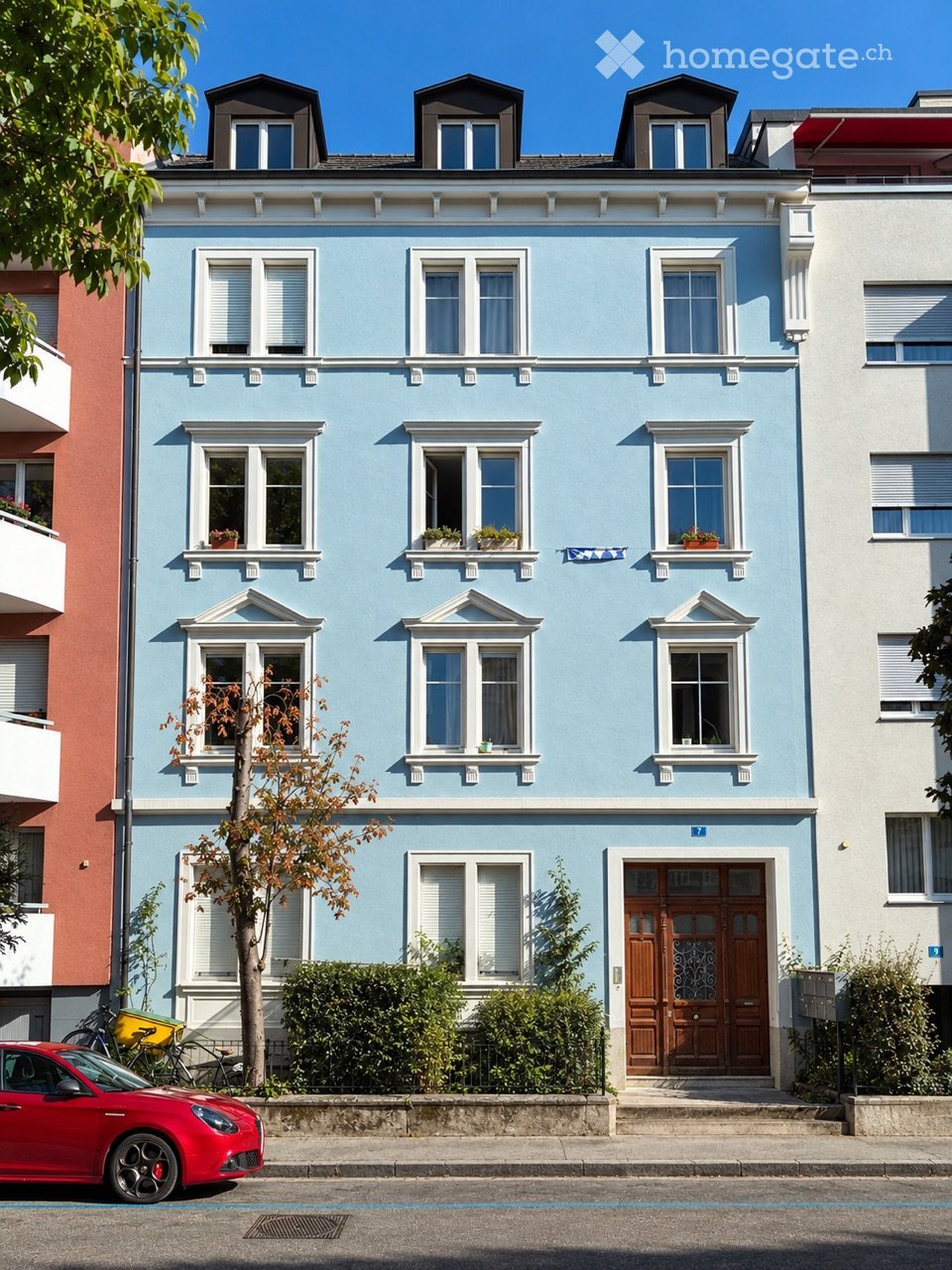 A three-story residential building with light blue facade, white window frames, decorative moldings, and small balconies with potted plants. The entrance features a wooden double door, and a red car is parked on the street in front.