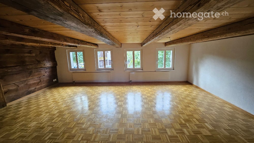 Empty room with wooden floor, exposed wooden beams on the ceiling, and four windows with white frames. The walls are plain and there are no furnishings or decorations.