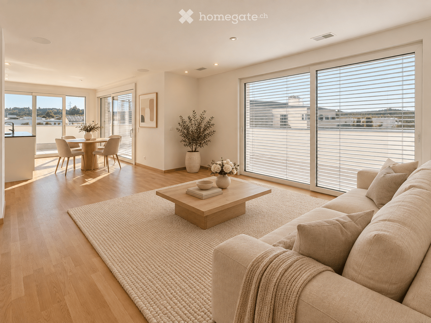 Modern living room with large windows and a balcony, featuring a beige sofa, wooden coffee table, dining area with chairs, and decorative vases.