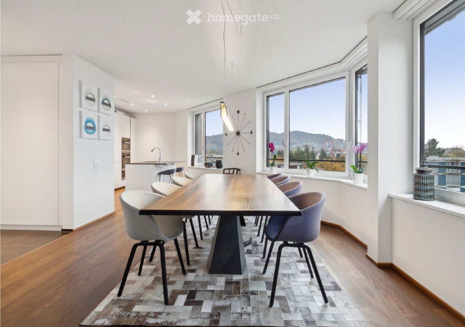Modern dining area with a long wooden table surrounded by contemporary chairs, a kitchen island with stools, large windows offering a scenic view, and decorative vases on the windowsill.