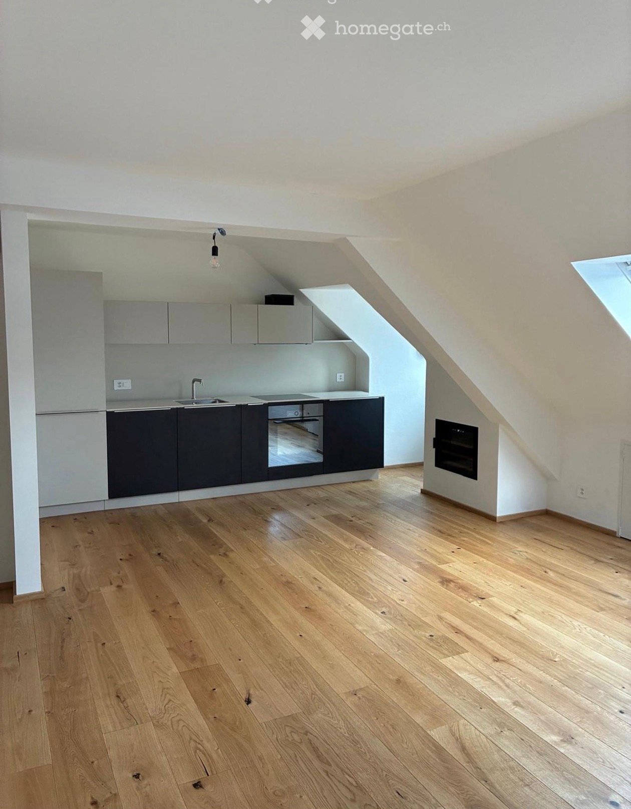 An empty kitchen with light wooden flooring, grey cabinets, and a built-in oven. The space features a modern design with a sink and a hanging light fixture.