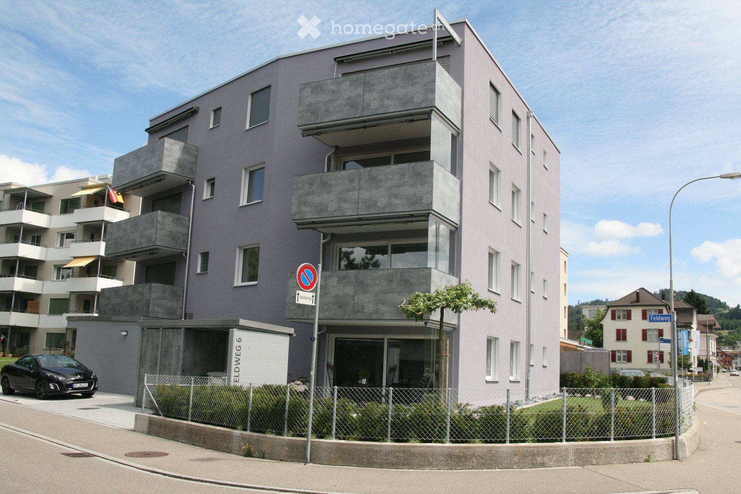 Modern multi-story apartment building with balconies, grey facade, and concrete elements. A car is parked in front, and a street sign is visible.