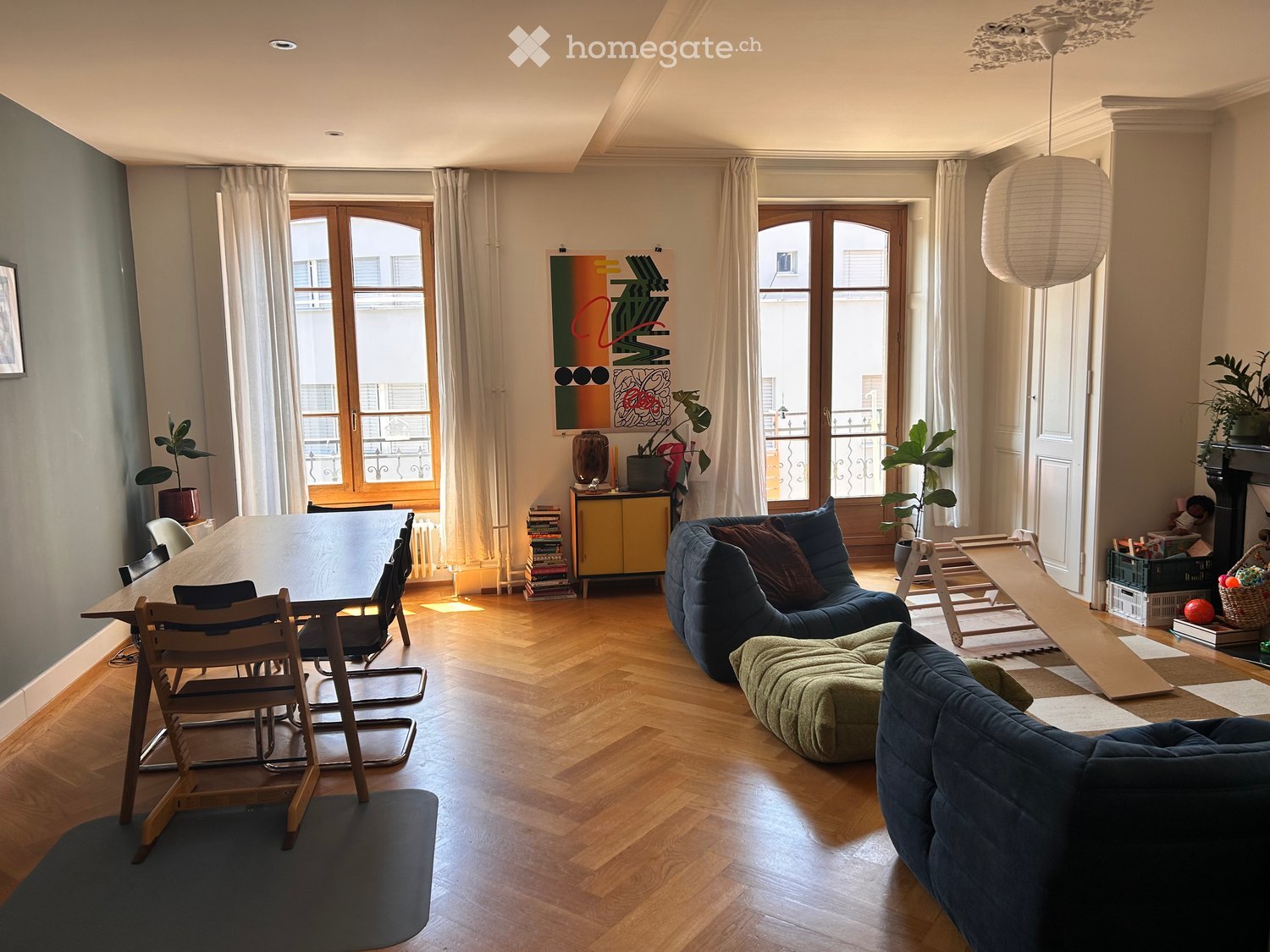 Living room with wooden flooring, large windows, sofa, ottoman, chairs, table, potted plants, and decorative items.