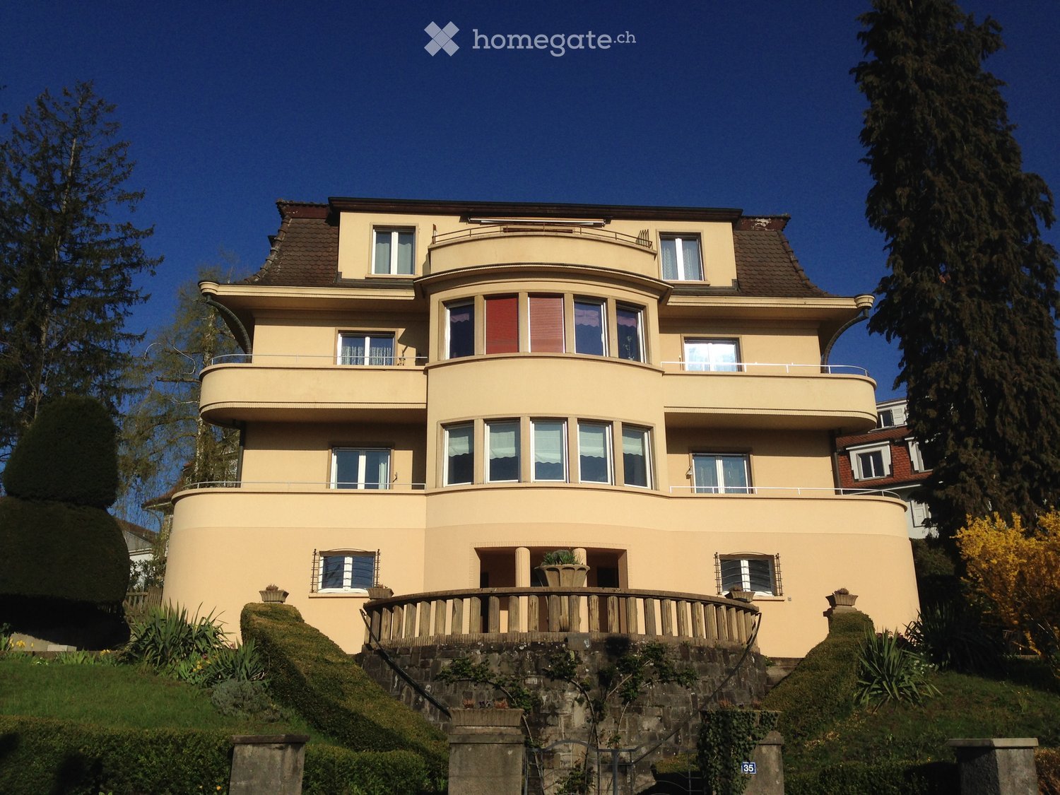A three-story house with a curved facade, multiple balconies, and large windows, situated on a raised ground floor with a stone staircase leading up to it.