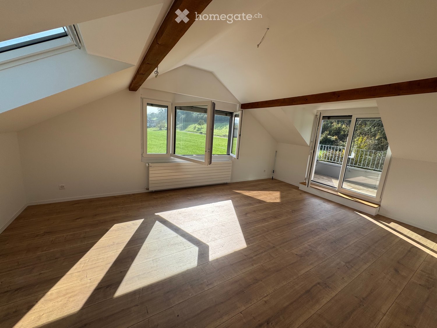 Bright, empty attic room with wooden flooring, large windows, and a sliding door leading to a balcony.