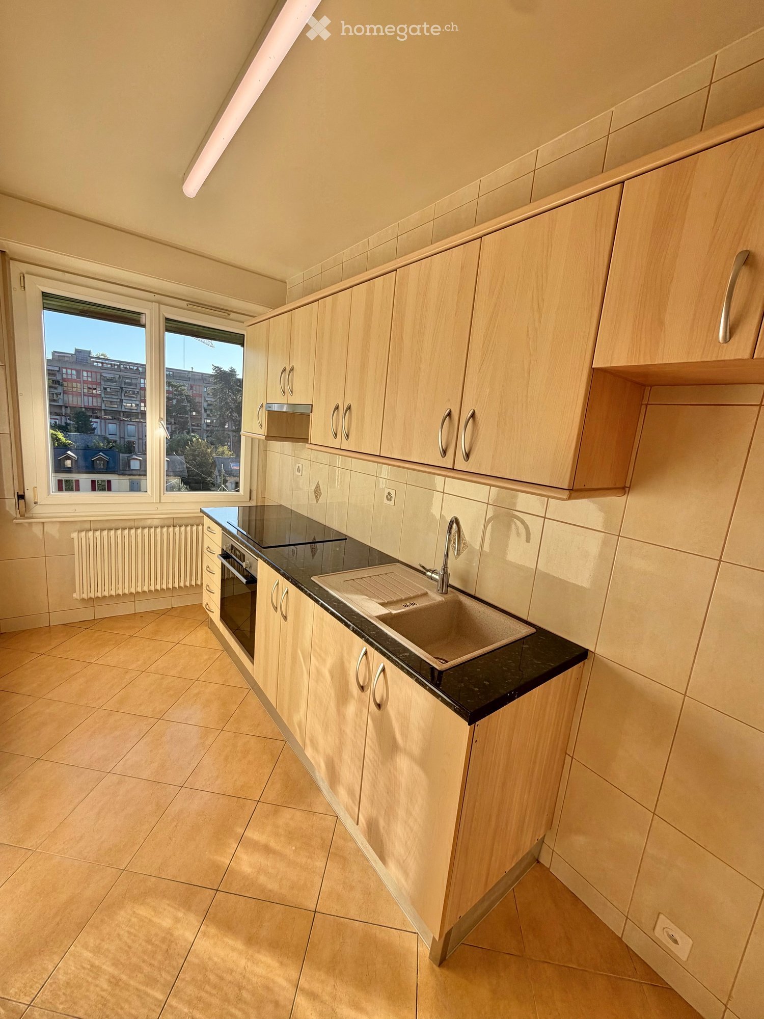 Modern kitchen with light wooden cabinets, black countertop, sink, and oven. Large windows provide natural light.