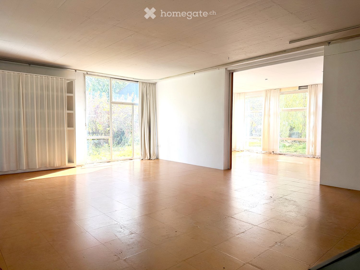 Empty room with wooden flooring, large windows with white curtains, and natural light flooding the space. No furniture or equipment present.
