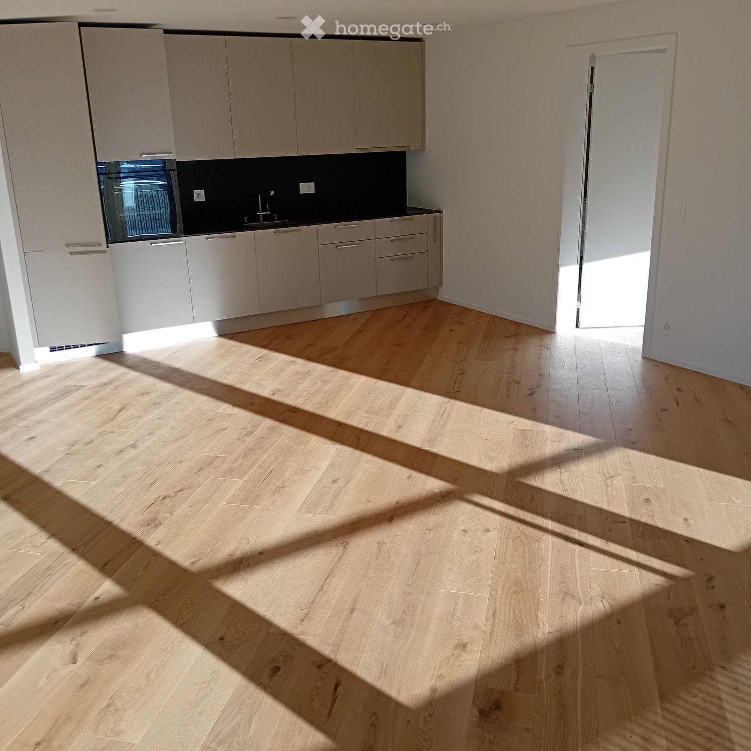 Empty kitchen with light wood flooring, modern cabinetry, and a built-in oven. Sunlight streams in from a window, casting shadows on the floor.