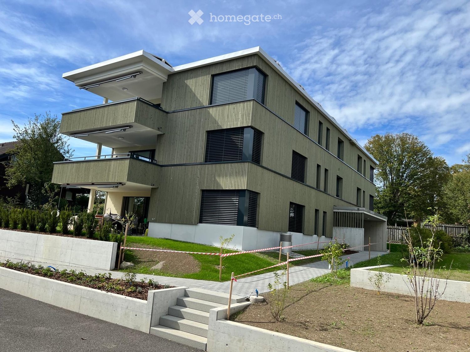 Modern two-story house with green siding, large windows, and balconies. The house features a raised ground floor, a well-maintained lawn, and a paved pathway leading to the entrance.