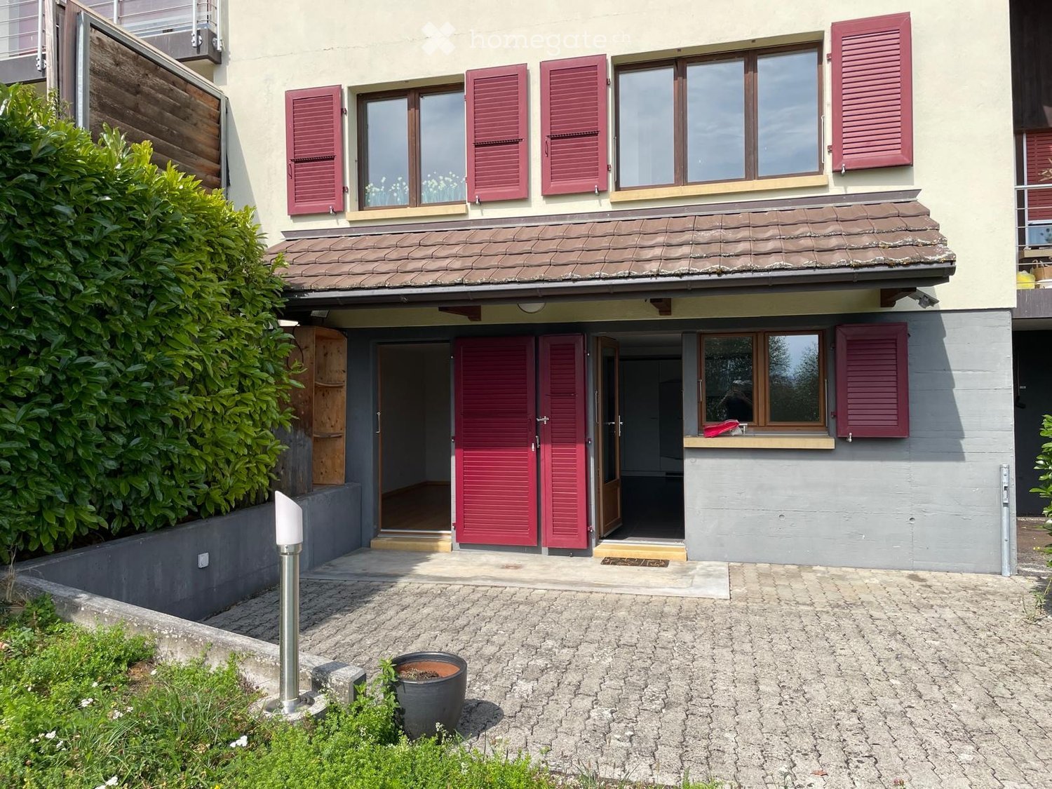 Two-story house with red shutters, beige walls, and a tiled roof. Features a raised ground floor, a small entrance porch, and a paved driveway.