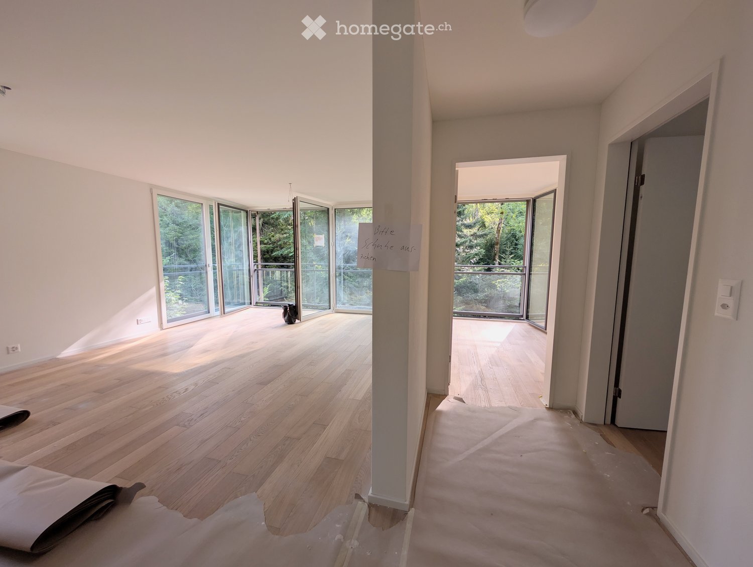 Empty hallway with wooden flooring, white walls, large glass doors leading to a balcony, and a small decorative item visible in the background.