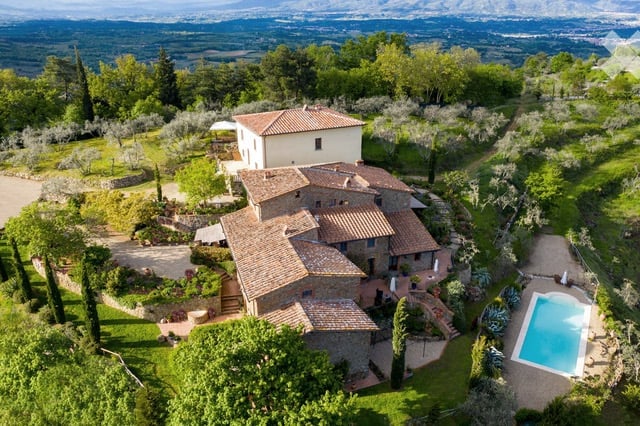 Aerial view of a large, rustic villa with terracotta roofs, surrounded by lush greenery, a swimming pool, and scenic countryside views.