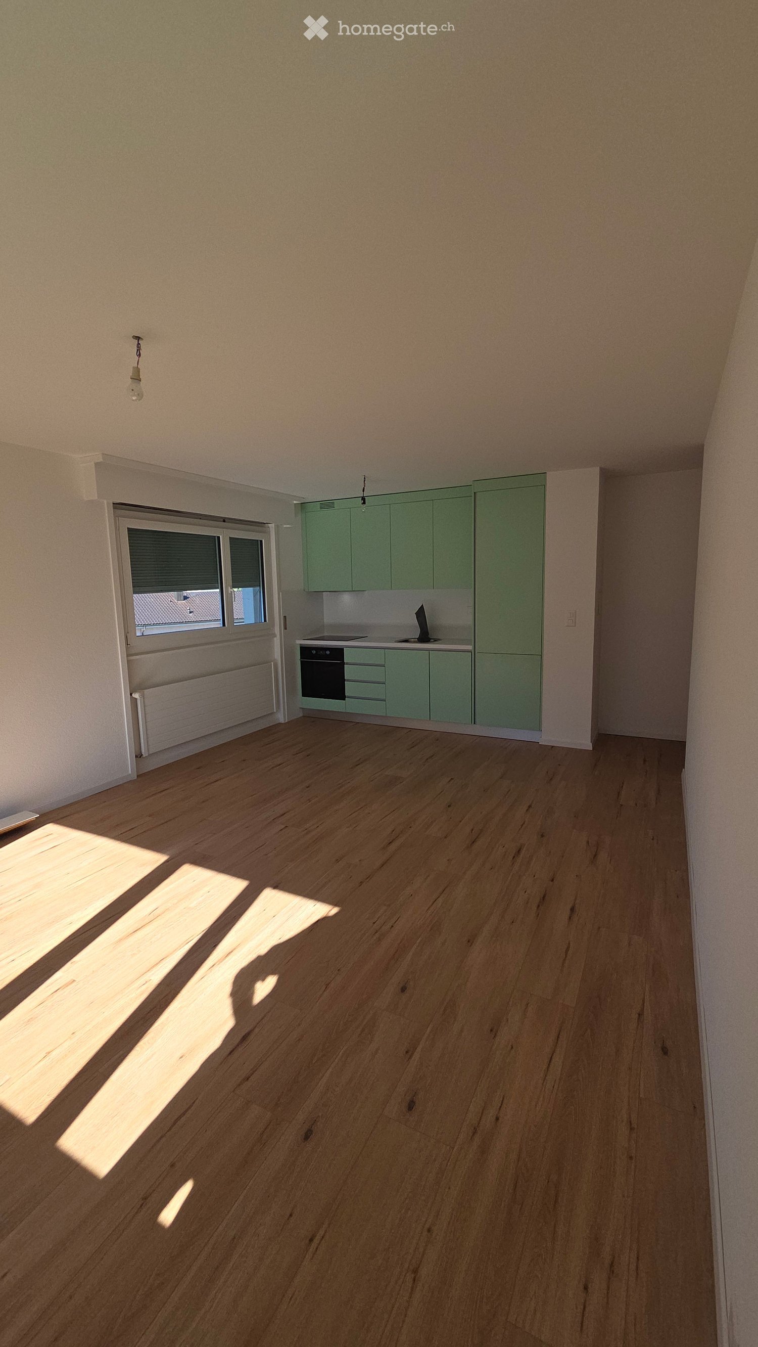 Empty kitchen with light green cabinets, wooden flooring, and a window with blinds. No furniture or appliances are present.