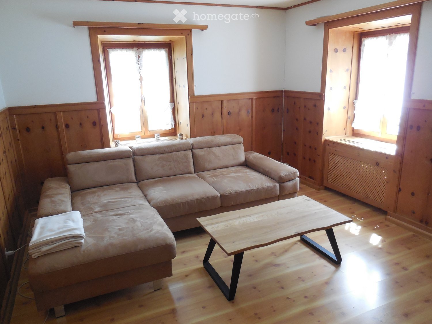 Living room with wooden walls, hardwood floor, large windows, beige sectional sofa, wooden coffee table, and folded blanket on sofa.