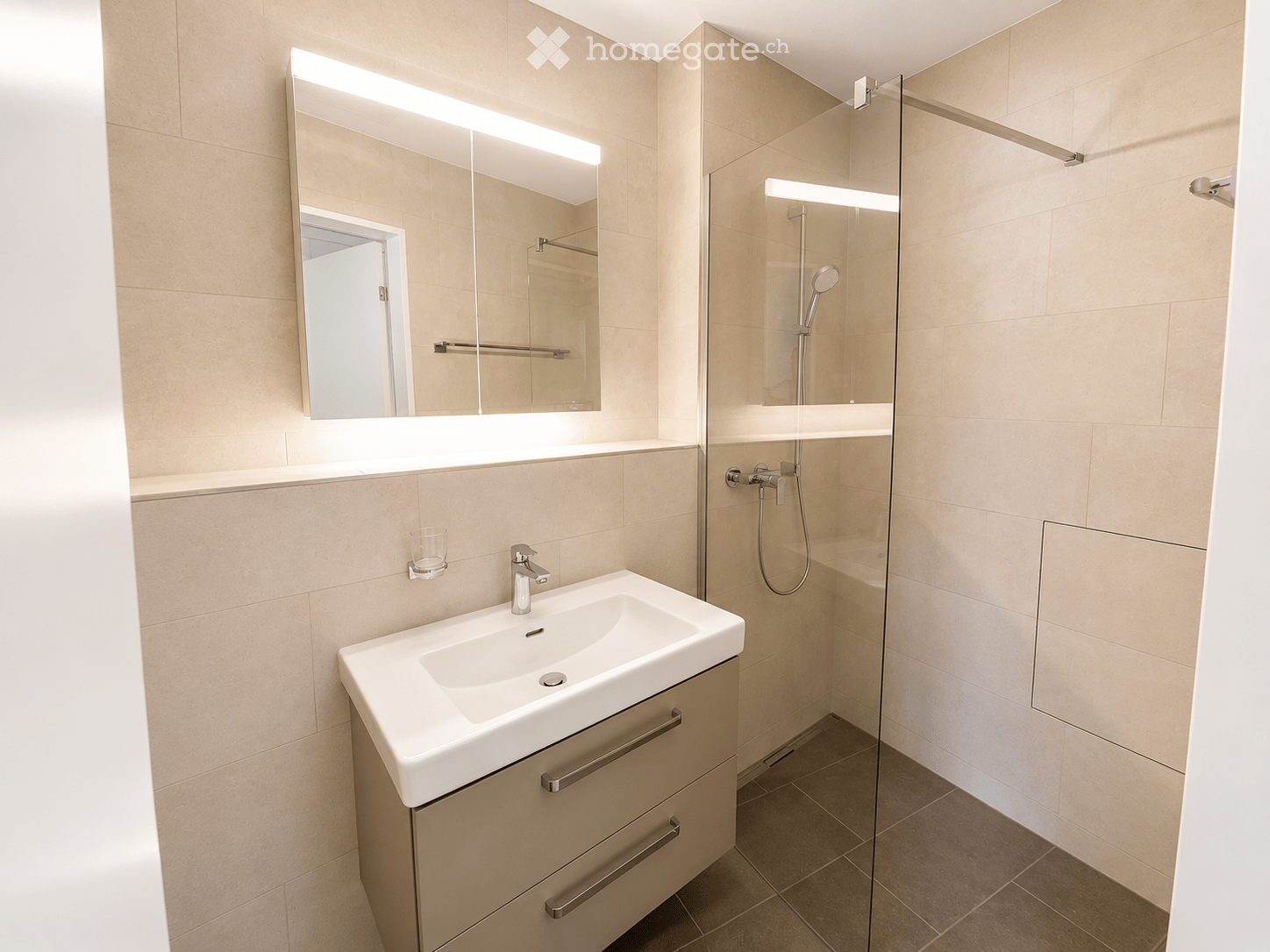 Modern bathroom with a white sink, mirror with built-in lighting, glass-enclosed shower, and a cup on the wall.
