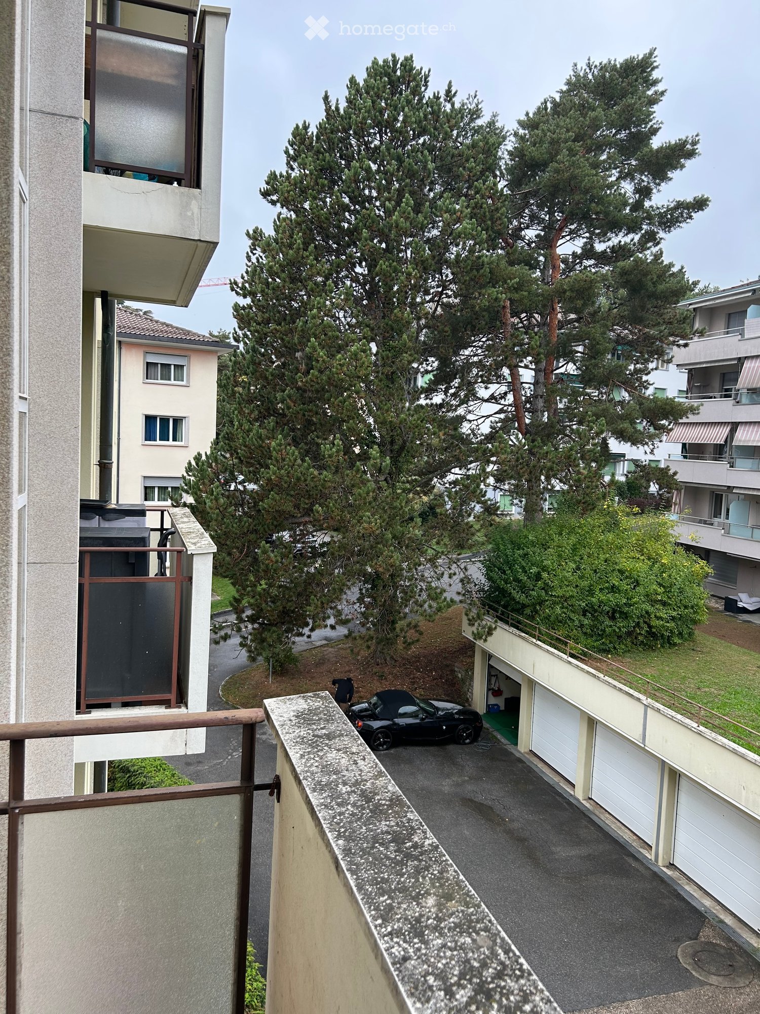 Balcony overlooking a parking area with a black car, surrounded by trees and neighboring buildings.