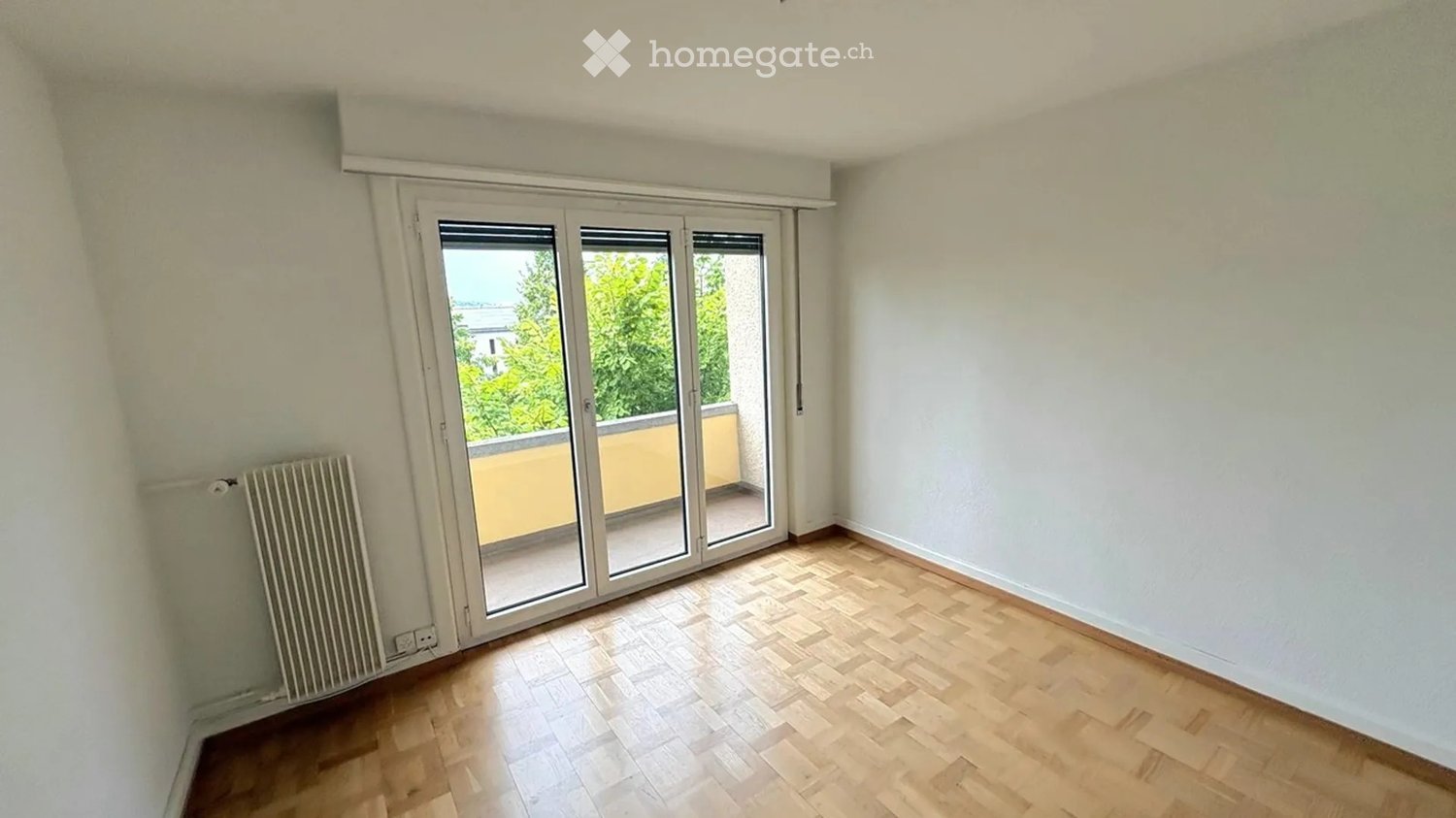Empty room with wooden flooring, white walls, large sliding glass doors leading to a balcony, radiator on the left side.