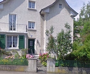 Two-story house with white facade, green shutters, and a small balcony on the upper floor. The house is surrounded by a low fence and some greenery, including bushes and small trees.