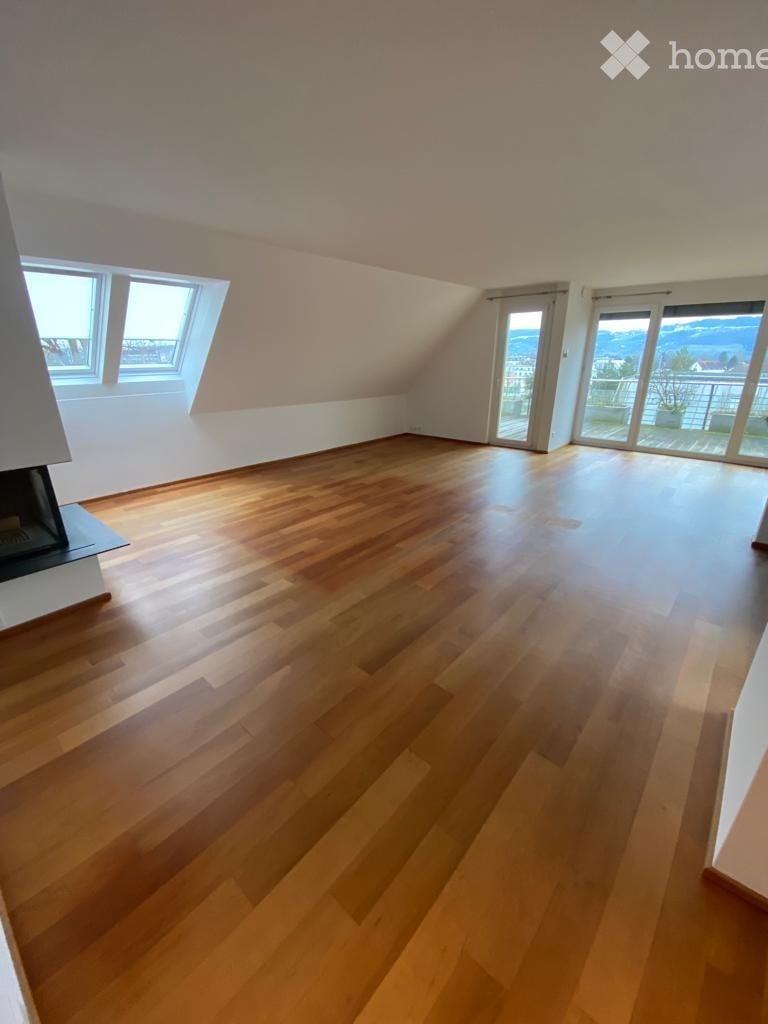 Bright, empty attic room with wooden flooring, large windows, and sliding glass doors leading to a balcony with a scenic view.