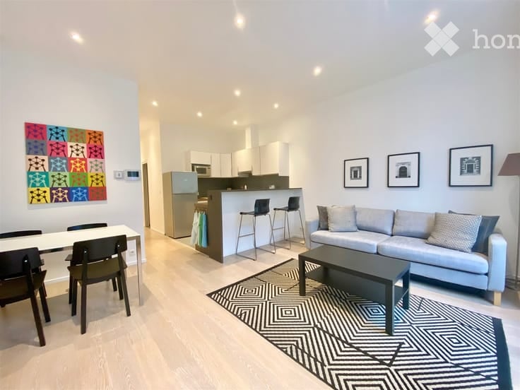 Modern open-plan living and dining area with a grey sofa, coffee table, dining table with chairs, kitchen island with stools, and a refrigerator. The space features a geometric rug, artwork on the walls, and recessed lighting.