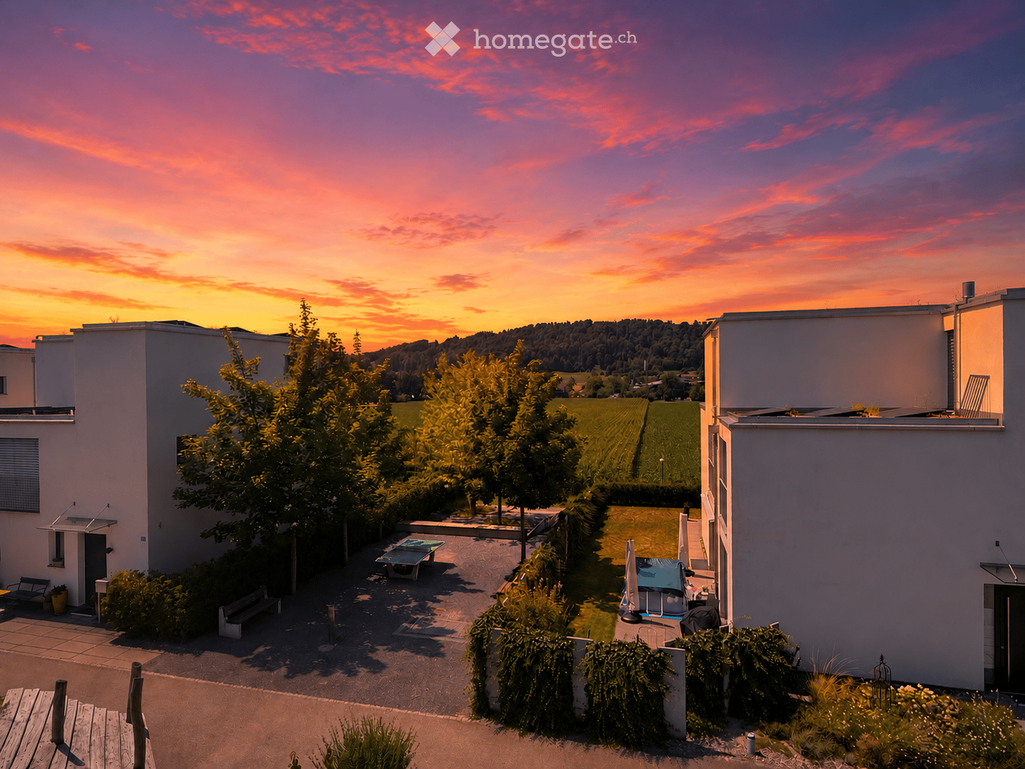 Courtyard with benches, a table, and an umbrella, surrounded by modern buildings and trees, with a scenic sunset view in the background.