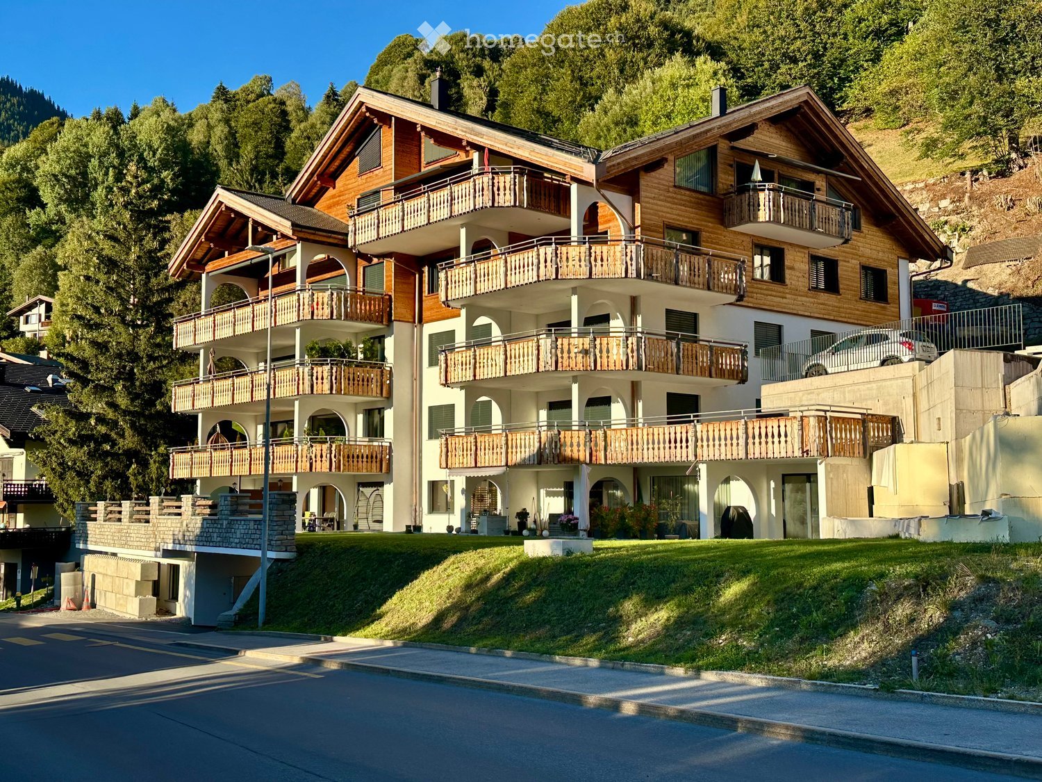 A large, multi-story wooden house with multiple balconies and a well-maintained lawn, situated on a hillside with trees in the background.