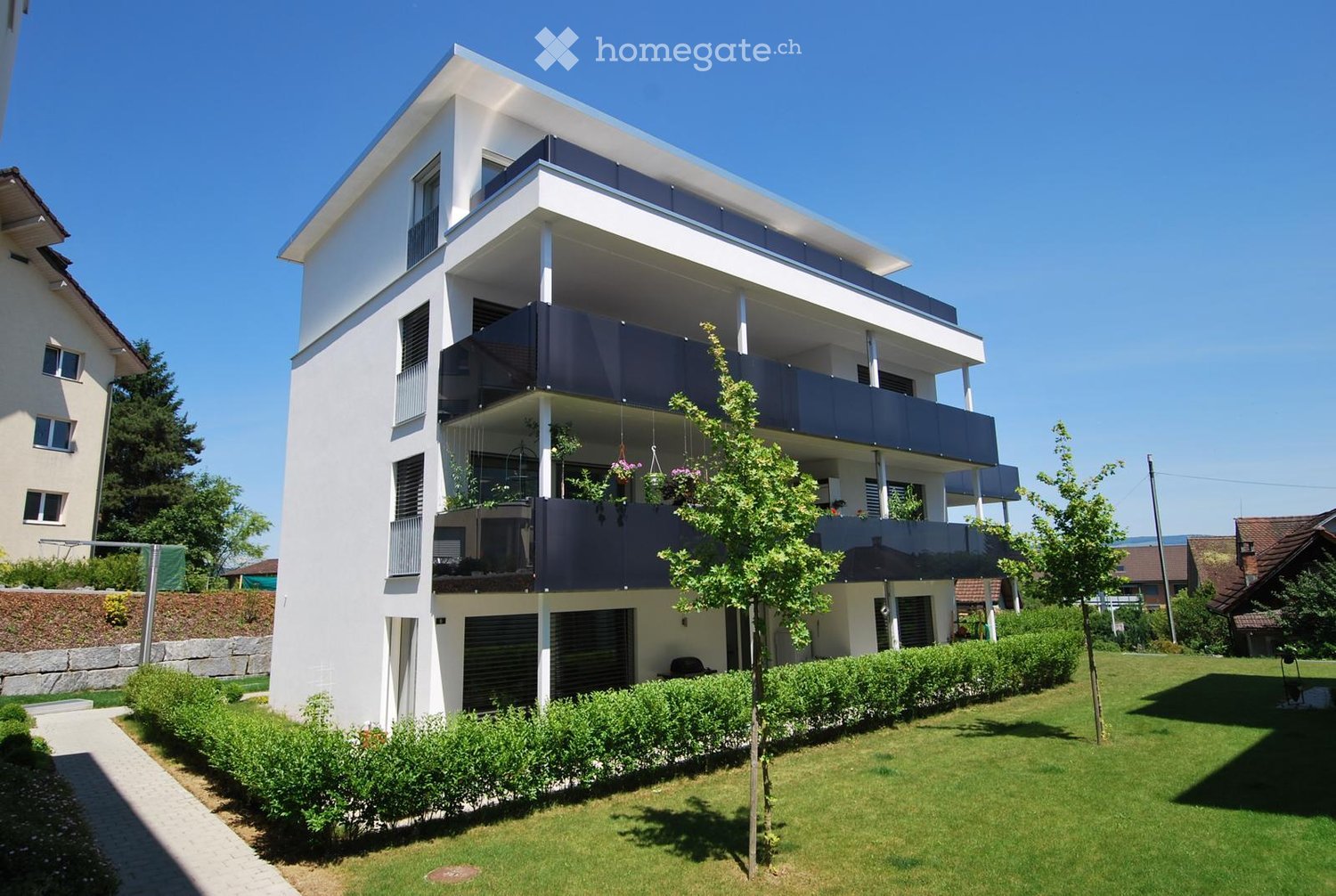 Modern two-story house with white exterior, large windows, balconies, and a well-maintained lawn with shrubs and a small tree.