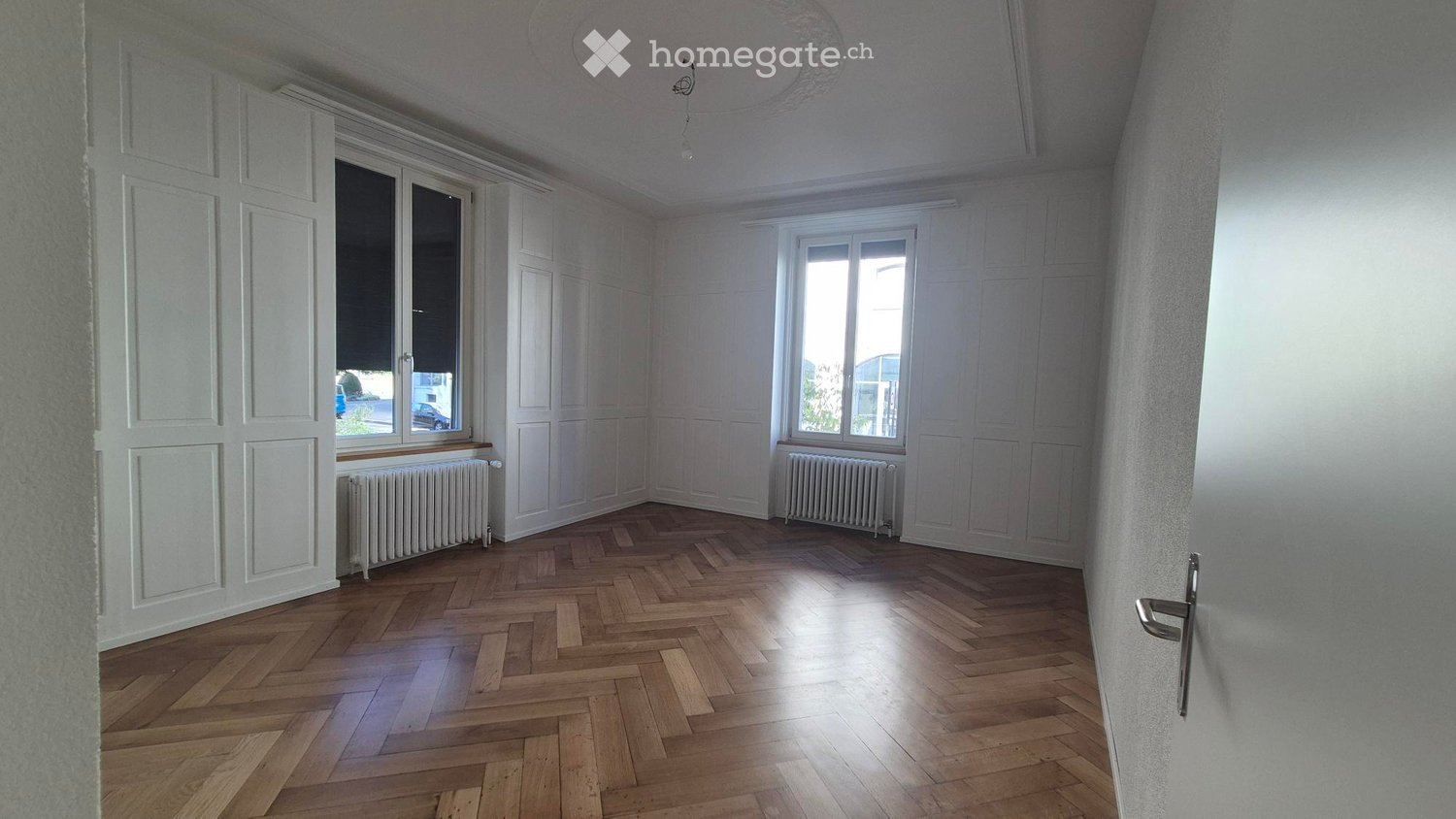 Empty room with wooden herringbone flooring, white walls, large windows, radiators, and a door.