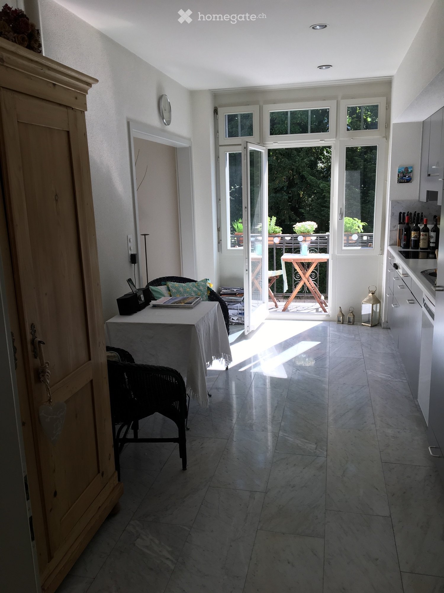 Bright, airy room with marble flooring, a small table with a cloth, a wicker chair, a wooden cabinet, and a view to a balcony with outdoor furniture.