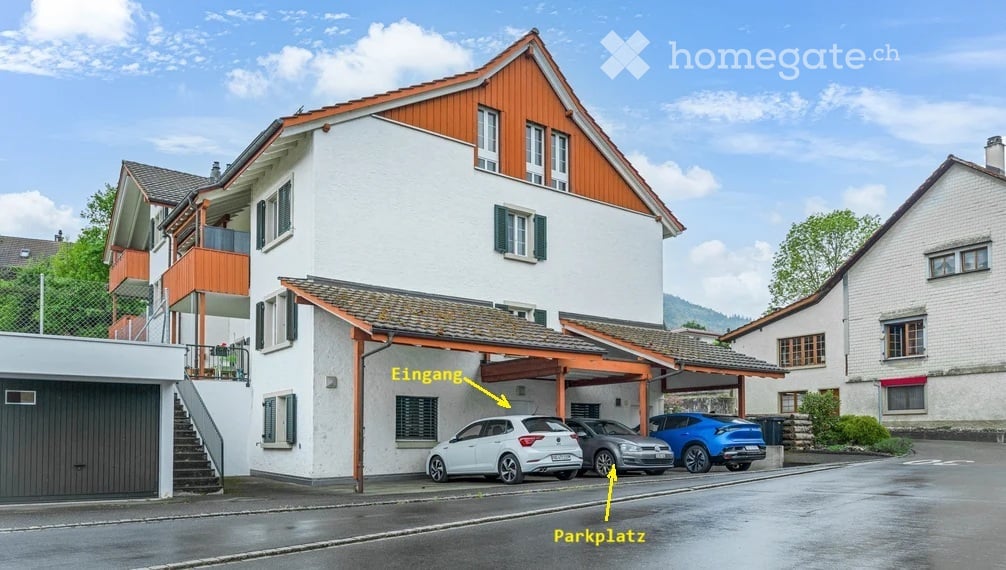 Two-story house with white walls, wooden roof, and green shutters. Features a covered entrance, garage, and parking area with several cars.