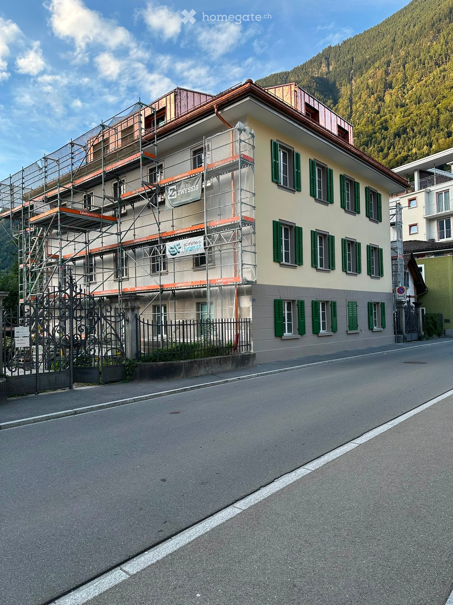 A multi-story building with green shutters and scaffolding on the upper floors, located on a street with a mountainous backdrop.