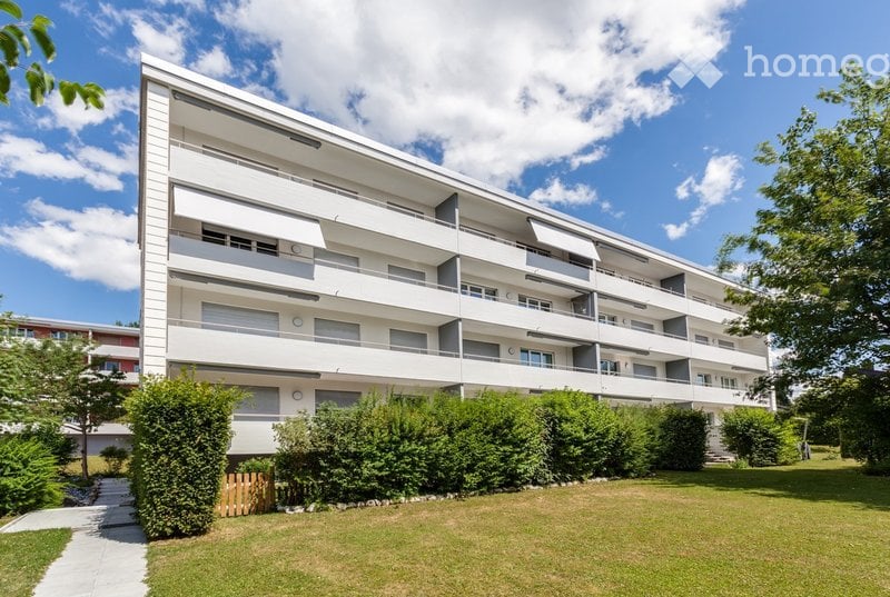 Modern multi-story apartment building with white facade, balconies, and surrounding greenery.