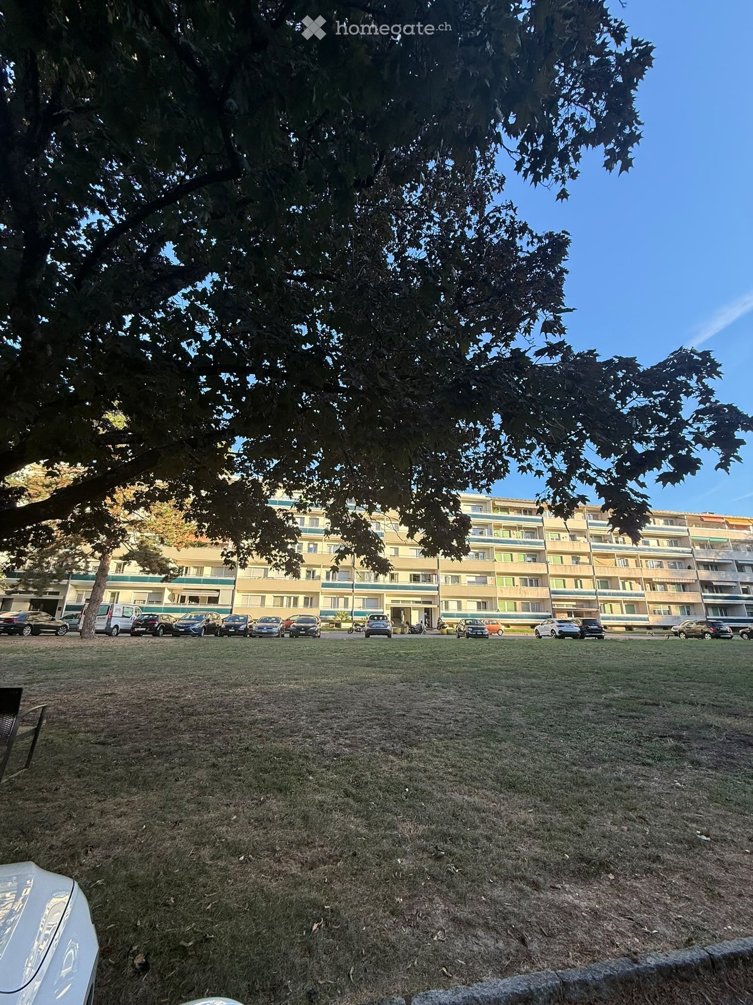 Multi-story residential building with balconies, surrounded by a grassy area with parked cars.