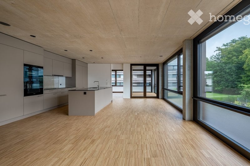 Open-plan kitchen and living area with light wood flooring, white cabinetry, and large windows offering a view of greenery. The space is unfurnished, with only built-in cabinetry and a kitchen island visible.