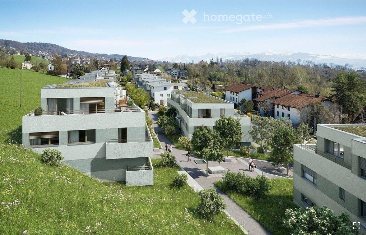 Aerial view of a modern residential complex with multiple buildings, green roofs, and surrounding greenery. The buildings are light-colored with large windows and balconies, situated on a hillside with a scenic backdrop of mountains and trees.