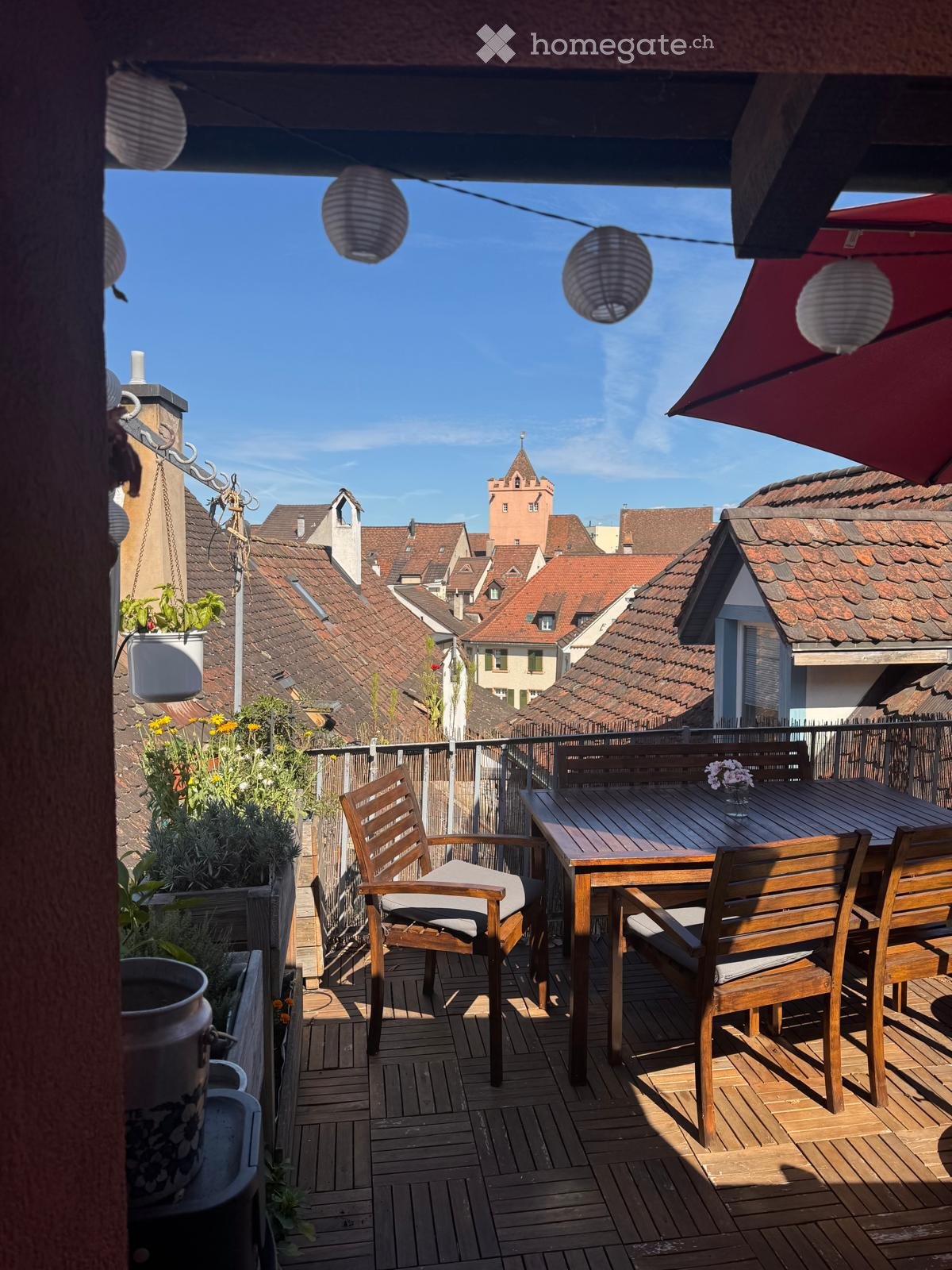 Wooden balcony with a table, chairs, and potted plants, overlooking a town with red-tiled roofs and a tower.